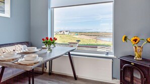 A dining area with a table set for four, a window with a view of a beach and buildings, and a vase with red flowers on the table. - 2 Bed in Amble (oc-l28057) (Amble)