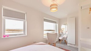 A bedroom with a large bed, two windows with a view of the beach, a chair, a vanity with a mirror, and a hanging light fixture. - 2 Bed in Amble (oc-l28057) (Amble)