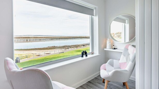 A room with a large window overlooking a pier and beach, two white chairs, a round mirror, and a small shelf with toiletries. - 2 Bed in Amble (oc-l28057) (Amble)