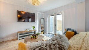 A modern living room with a flat-screen TV, a round coffee table with fruit, a sofa with a fluffy throw, and a large mirror. - 1 Bed in Newquay (oc-l28083) (Newquay)