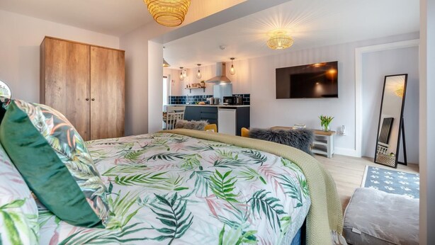 A modern bedroom with a large bed, a wooden wardrobe, a kitchen in the background, and a TV mounted on the wall. - 1 Bed in Newquay (oc-l28083) (Newquay)