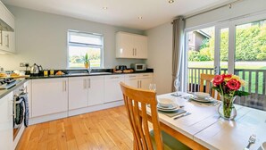 A modern kitchen with white cabinets, a wooden dining table with chairs, and a view of a garden through large windows. - 2 Bed in Winnards Perch (oc-w28086) (Winnard's Perch)