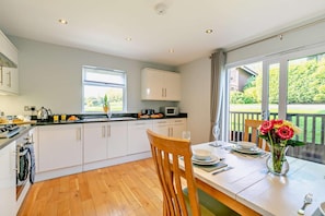A modern kitchen with white cabinets, a wooden dining table with chairs, and a view of a garden through large windows. - 2 Bed in Winnards Perch (oc-w28086) (Winnard's Perch)