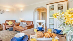 A living room with a brown leather sofa, a dining table with breakfast items, and a kitchen area with a cabinet. - 2 Bed in Putsborough (oc-a22903) (Croyde)