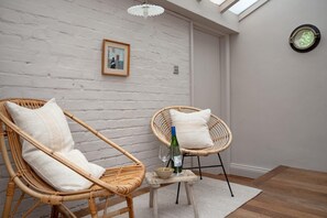 Ein Zimmer mit zwei Korbstühlen, einem kleinen Tisch mit Flasche und Glas sowie einem gerahmten Bild an der Wand.