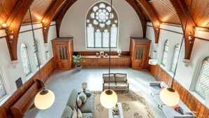 A spacious loft with high ceilings, exposed wooden beams, and a large arched window. Furnishings include a sofa, armchairs, and a coffee table. - 3 Bed in Yoxford (oc-t28018) (Aldeburgh)