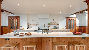 A spacious kitchen with a central island, wooden bar stools, and a large window with stained glass. - 3 Bed in Yoxford (oc-t28018) (Aldeburgh)
