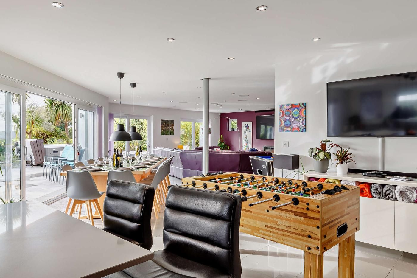 A modern living room with a foosball table, a dining area with chairs, and a television on the wall.