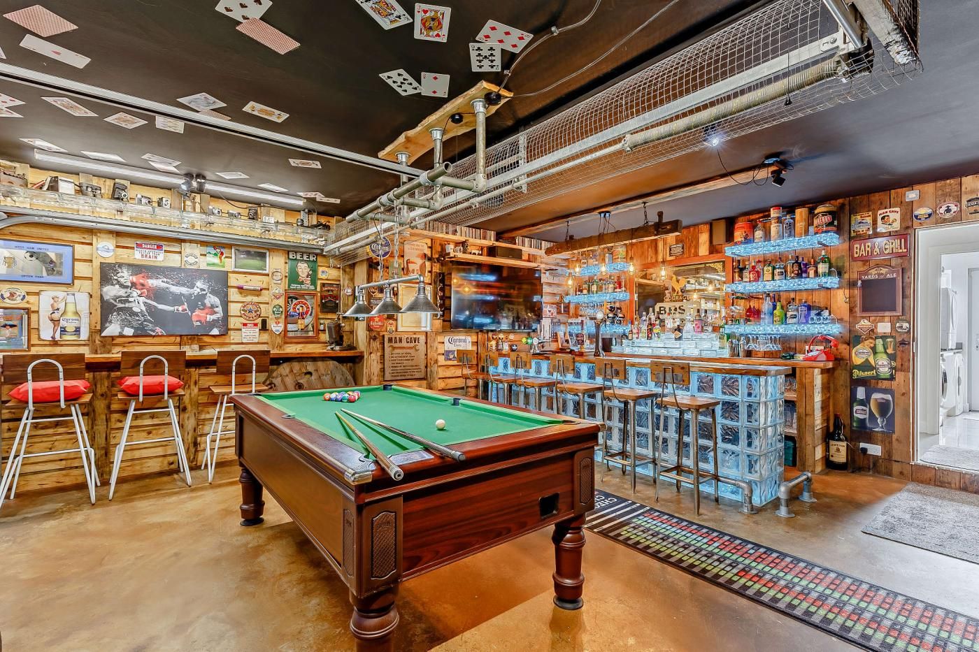 Zona de bar con mesa de billar, taburetes y estantes llenos de botellas.