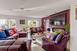 A modern living room with leather sofas, a flat-screen TV, and a large window. - 5 Bed in Tavistock (oc-m28107) (Tavistock)