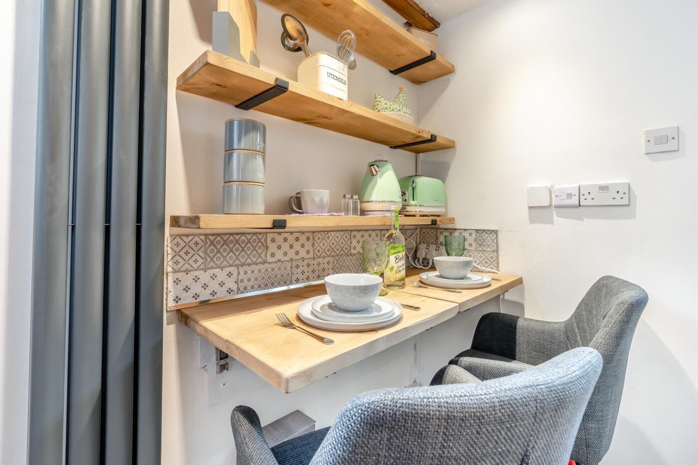 Une cuisine avec des étagères en bois, une bouilloire, une bouilloire verte, une bouteille et une chaise.
