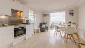 Una cocina moderna con armarios blancos, un microondas integrado y un horno. Hay un comedor con sillas y una mesa blancos, y una vista al mar a través de una puerta de cristal.