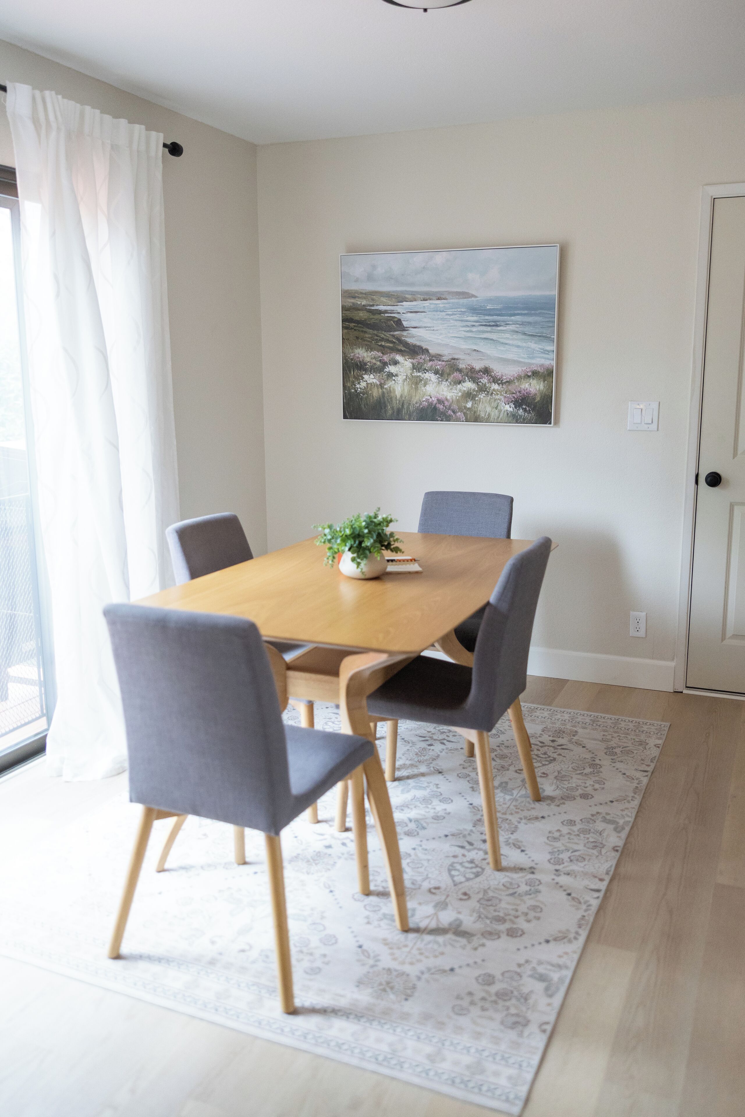 A dining area with a wooden table, grey chairs, a potted plant, and a painting of a landscape on the wall.