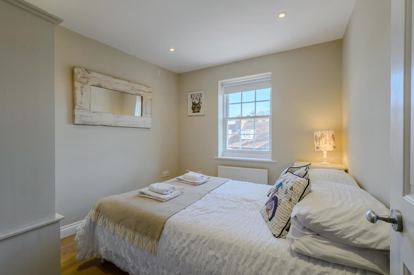 A bedroom with a bed, a window, a mirror, and a bedside table with a lamp.