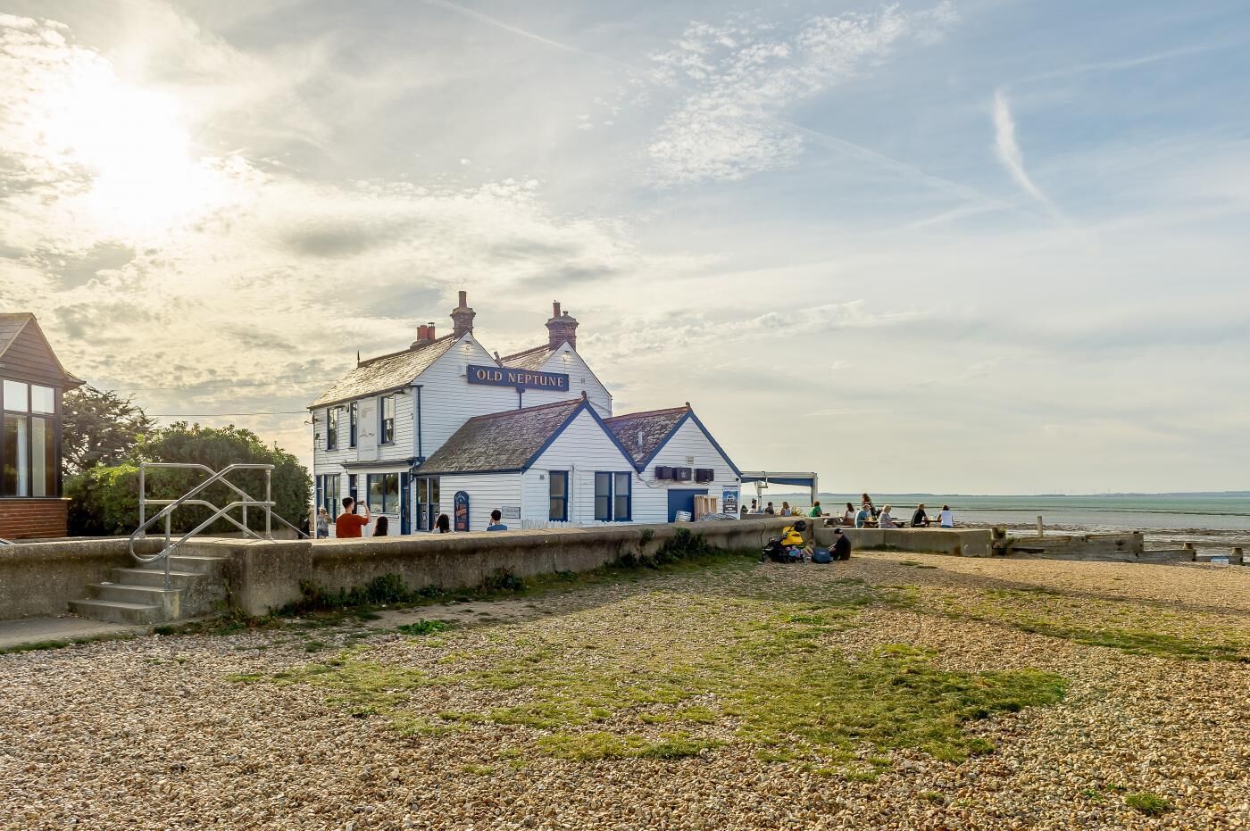 Un bâtiment blanc, sur lequel on peut lire l’inscription « Old Neptune », se dresse sur une plage de galets, avec des personnes en arrière-plan.