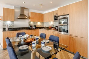 A modern kitchen with wooden cabinets, a glass dining table, and a built-in oven. - 3 Bed in Torquay (oc-a28109) (Torquay)