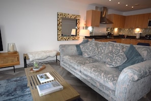 A modern living room with a patterned sofa, a wooden coffee table with books, a small side table with a lamp, and a kitchen in the background. - 3 Bed in Torquay (oc-a28109) (Torquay)