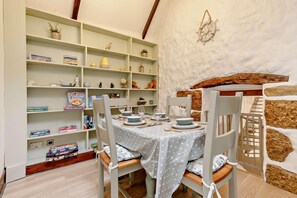 Un coin repas avec une table dressée pour quatre personnes, entourée de chaises blanches dotées de coussins gris. Une bibliothèque remplie de divers objets se trouve à gauche.