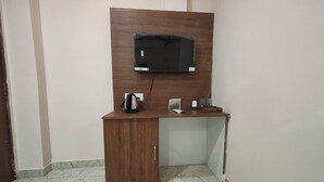 A small room with a wooden desk, a black flat-screen TV mounted on the wall, a kettle, a small white device, and a glass container. - HOTEL ARDENT (Ranchi)