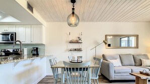 Eine moderne Küche mit weißen Schränken, ein Essbereich mit rundem Tisch und Stühlen sowie ein Wohnzimmer mit Sofa und Couchtisch.