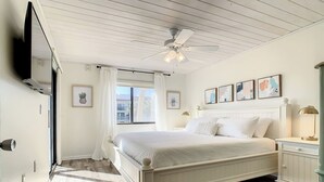 Ein Schlafzimmer mit Bett, Nachttisch, Deckenventilator, Fenster mit Vorhängen und gerahmten Bildern an der Wand.