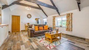 A spacious loft with exposed wooden beams, a cozy seating area with a sofa and cushions, a dining table, and a kitchen area. - 1 Bed in St. Tudy (oc-t28171) (Michaelstow)
