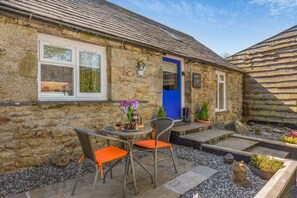 Un bâtiment en pierre avec une porte bleue, un petit espace extérieur aménagé avec des chaises en osier et une table, et un jardin avec des plantes en pot.