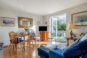 A spacious living room with a blue sofa, wooden chairs, a dining table with breakfast items, a TV, and a balcony with a garden view. - 1 Bed in Long Melford (oc-s28172) (Long Melford)