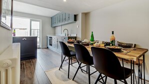 A modern kitchen with grey cabinets, a dining table with black chairs, and a view of the city through a large window. - 3 Bed in Southwold (oc-r11173) (Southwold)
