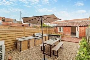 An outdoor patio with a grill, wooden table, and benches. - 3 Bed in Southwold (oc-r11173) (Southwold)