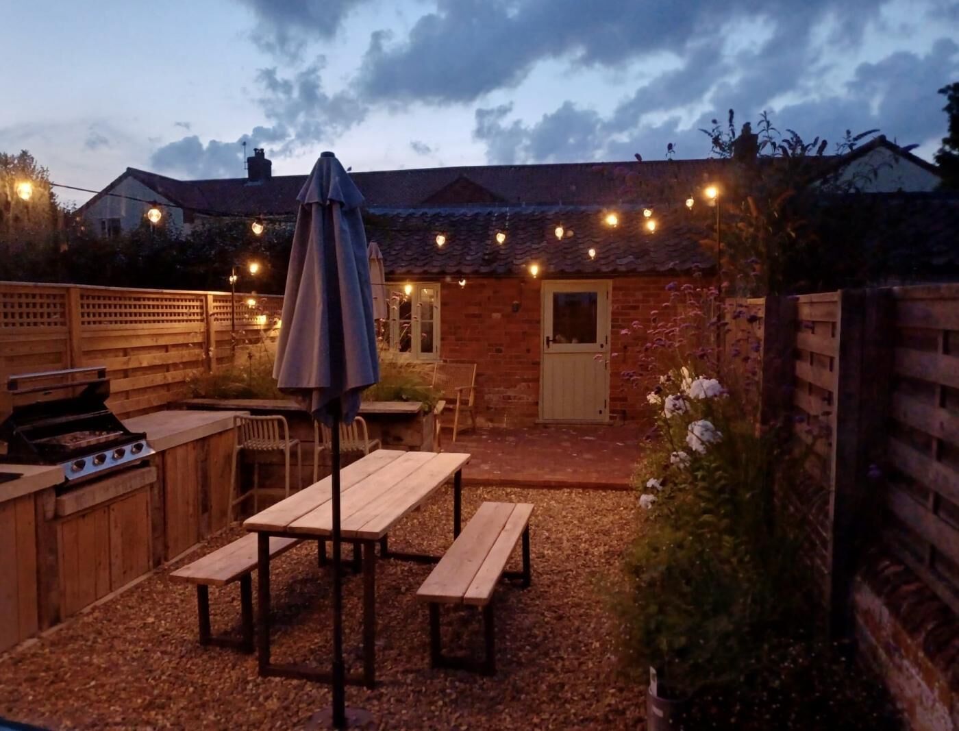 En udendørs patio med træborde og bænke, grill, lyskæder og en murstensbygning i baggrunden.