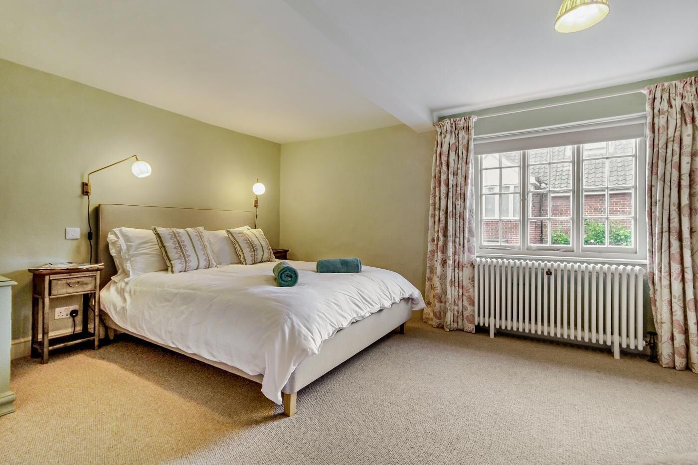 A bedroom with a bed, nightstands, a lamp, a window with curtains, and a radiator.