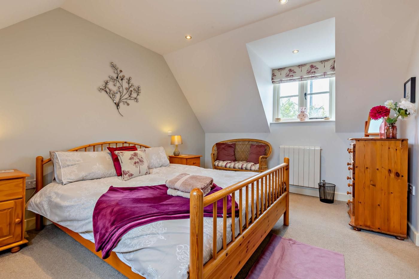 A bedroom with a sloped ceiling, a bed with a wooden frame, a wicker chair, a wooden cabinet, and a window with a view of greenery.