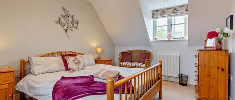 A bedroom with a sloped ceiling, a bed with a wooden frame, a wicker chair, a wooden cabinet, and a window with a view of greenery.