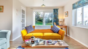 A bright living room with a yellow sofa, a coffee table with a tray of coffee and cups, and a view of a garden through large windows. - 4 Bed in West Bexington (oc-s28233) (West Bexington)
