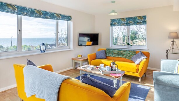 A bright living room with yellow sofas, a coffee table, and large windows overlooking the sea. - 4 Bed in West Bexington (oc-s28233) (West Bexington)