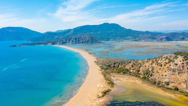 An aerial view of a coastal landscape with a sandy beach, clear blue waters, and distant mountains.