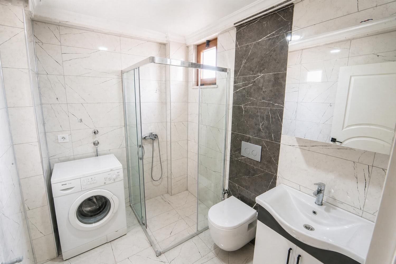 A bathroom with a glass-enclosed shower, a washing machine, a toilet, and a sink.