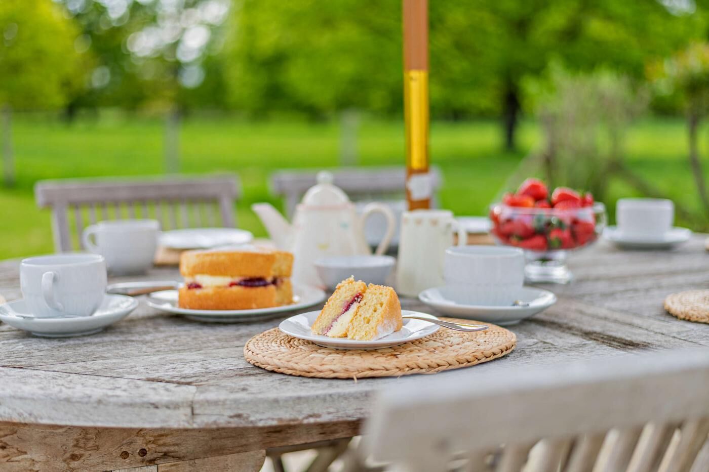 Una mesa rústica al aire libre con un montaje para picnic que incluye sándwiches, tazas, una tetera y un bol de fresas.