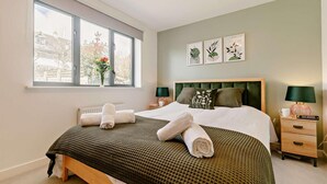 A bedroom with a large window, a bed with green bedding, two bedside tables with lamps, and three wall-mounted art pieces. - 2 Bed in Charlestown (oc-91449) (Charlestown)