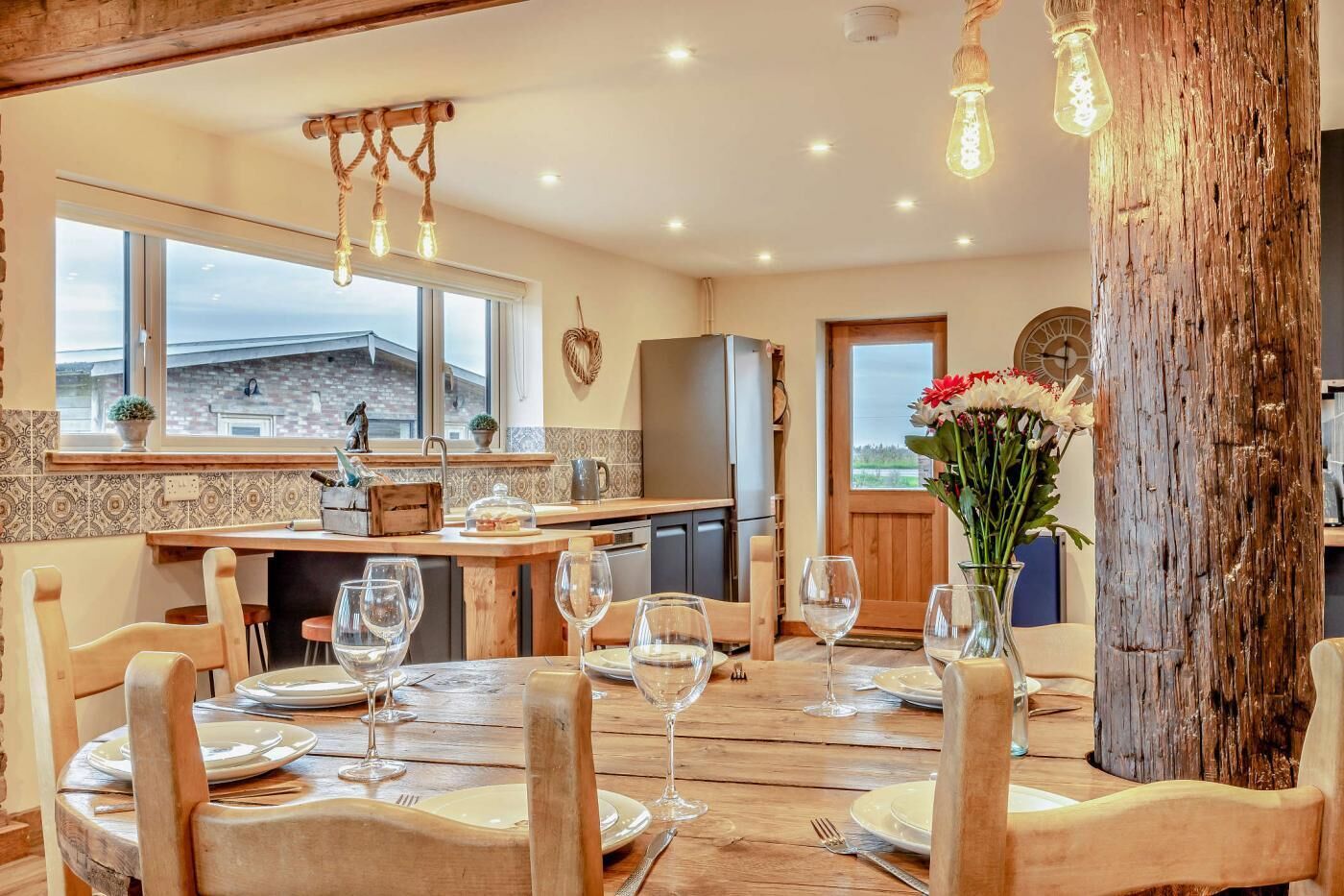 Een eetgedeelte met houten stoelen en een tafel voor zes personen, een keuken met een groot raam, en een rustieke houten zuil.