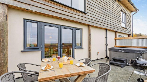 A wooden table with chairs and breakfast items outside a house with large windows and a barbecue grill. - 2 Bed in Outwell (oc-a28259) (Upwell)