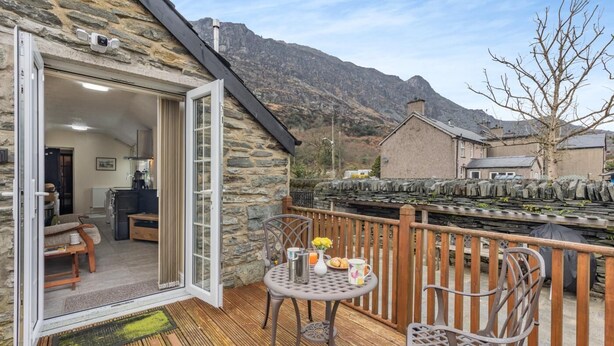 Un bâtiment en pierre doté d’une terrasse en bois, d’une table avec des chaises, et avec une chaîne de montagnes en arrière-plan.