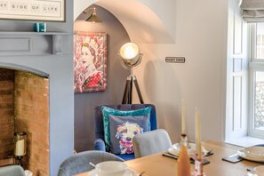 A dining area with a table set for two, a blue sofa with a floral cushion, a framed artwork above the sofa, a wall-mounted lamp, and a sign reading 'NAUGHTY CORNER'. - 4 Bed in East Rudham (oc-t28312) (East Rudham)