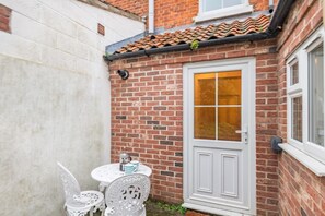A small, enclosed patio with a white door, a brick wall, and a tiled roof. There is a small table with chairs and a cup set on the table. - 2 Bed in East Runton (oc-m837) (East Runton)