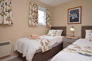 Ein Schlafzimmer mit zwei Betten, einem Fenster mit Vorhängen, einer Nachttischlampe und einem gerahmten Bild an der Wand.