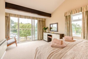 A bedroom with a large bed, a desk, and a chair. There are two windows with curtains, and a balcony is visible through the window.