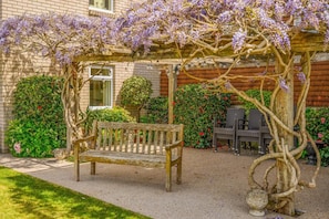 A garden with a wooden bench, a pergola with purple flowers, and a brick building in the background. - 3 Bed in Christchurch (oc-r27898) (Christchurch)