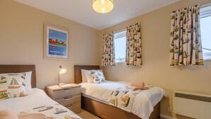Camera d'albergo con due letti, un comodino, una scrivania, una lampada, un quadro incorniciato e tende a motivi decorativi.