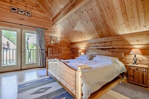 A wooden cabin bedroom with a bed, nightstands, and a window with a view of a house. - 3 Bed in Little Petherick (oc-c28311) (Little Petherick)